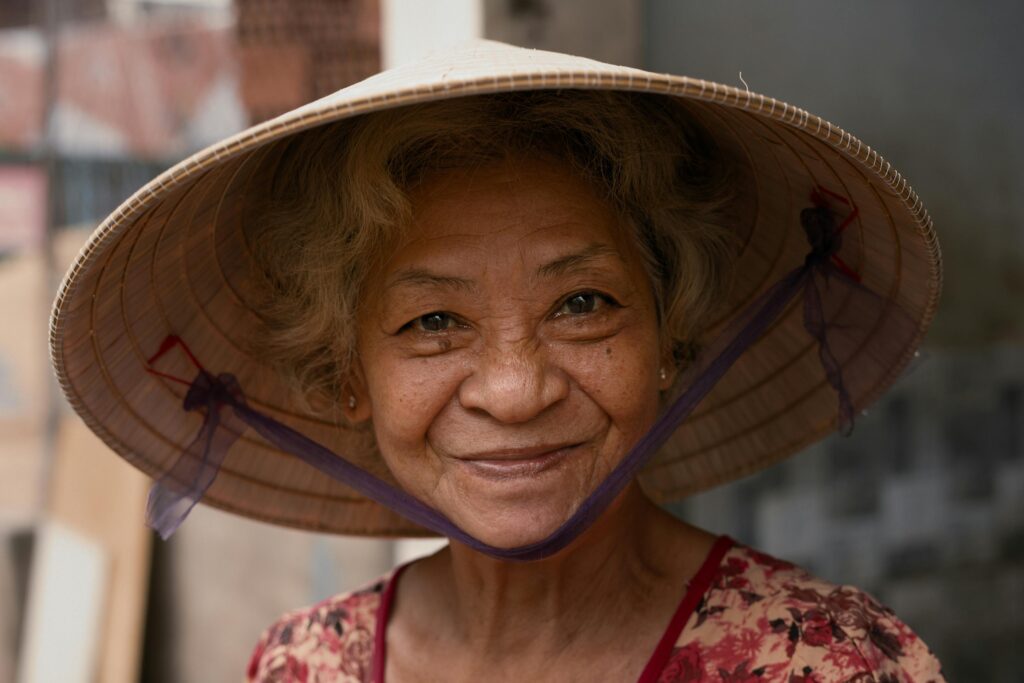 A cheerful elderly woman in a traditional conical hat smiles warmly in an outdoor urban setting.