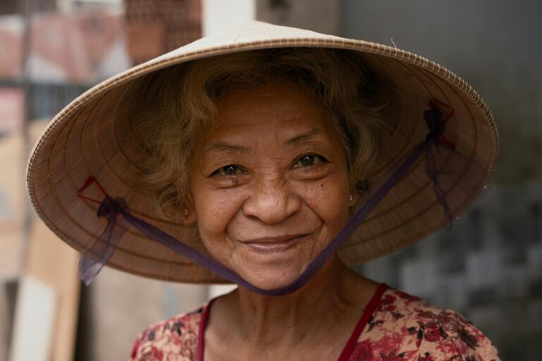A cheerful elderly woman in a traditional conical hat smiles warmly in an outdoor urban setting.